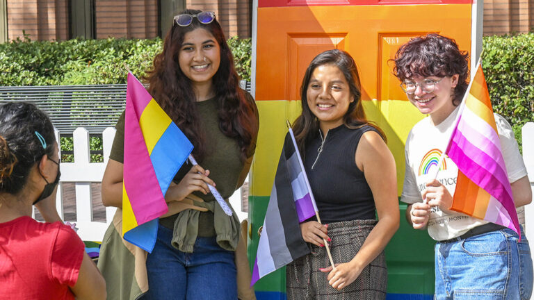 Three students pose in front of an rainbow colored door with LGBTQ+ flags.