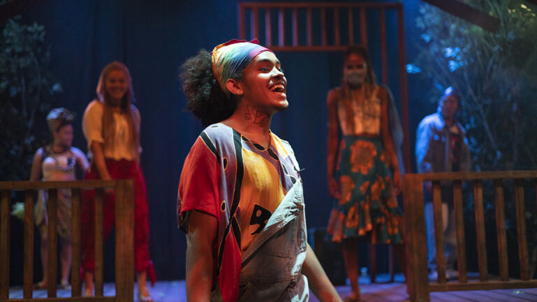 An actor singing, wearing a bandana and overalls, in a USC School of Dramatic Arts stage performance of 