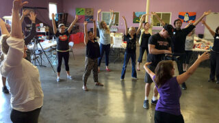 12 adults raising their hands as part of a dance exercise in a middle of an art studio