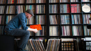 A man looking at a vinyl record with a wall of vinyl records behind him