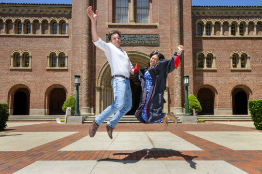 USC Thornton grads Heeseung Lee, right, and Victor Martinez Sara jump for a photograph in front of Bovard