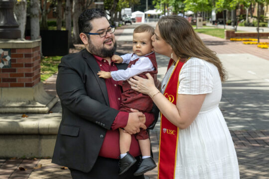 a graduate and her partner kissing their child
